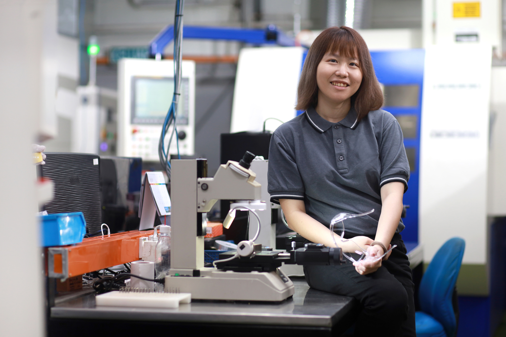 Portrait of smiling Asian female engineer sitting beside microscope in engineering factory